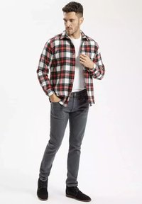 Czerwona i czarna flanelowa koszula w kratę na białej koszulce, w połączeniu z szarymi spodniami i czarnymi zamszowymi botkami. Model stoi w neutralnej pozycji.
