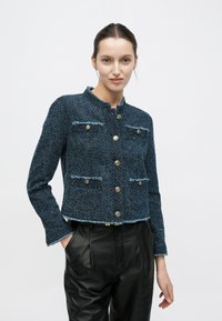 Giacca blu testurizzata con design corto, orlo sfrangiato, quattro bottoni e due tasche sul petto. Abbinata a pantaloni in pelle neri.