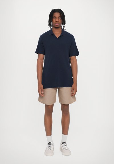 Polo de marina, de manga corta, corte relajado. Pantalones cortos beige, hasta la rodilla, de textura lisa. Calcetines blancos y zapatillas ligeras completan el conjunto.