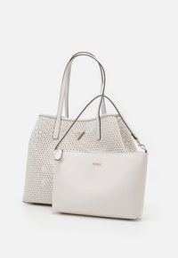 Deux sacs à main Guess couleur crème, l'un grand avec des perforations en forme de diamant et le logo triangle, l'autre plus petit avec une bandoulière simple et le nom de la marque.