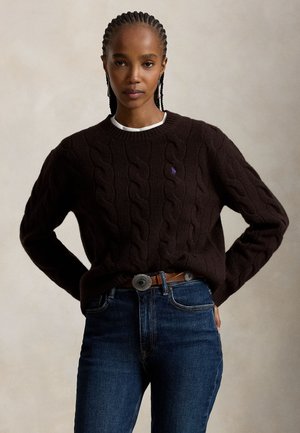 Femme aux cheveux tressés portant un pull en maille torsadée marron foncé et un jean bleu avec une ceinture marron, debout les mains sur les hanches.
