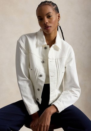 Femme aux cheveux tressés portant une veste en denim blanche boutonnée et un pantalon sombre, assise contre un fond neutre avec une posture décontractée.