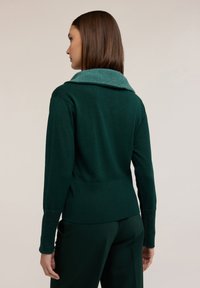 Donkergroene trui met een gestructureerde teal kleur kraag, lange mouwen en geribde manchetten. De achterkant heeft een aansluitend ontwerp en een lichte uitlopende vorm aan de zoom.