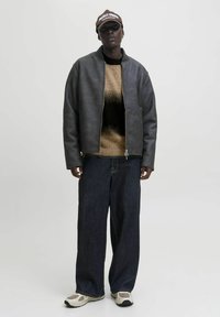 Person, die eine schwarze Lederjacke, einen braun-schwarzen Verlaufspullover, weite dunkle Jeans, beige Sneaker und eine braune Mütze mit weißem Text trägt.