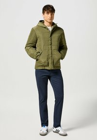 Chaqueta de plumas verde oliva con capucha, diseño acolchado, bolsillos frontales, combinada con pantalones azul marino y zapatillas blancas con acentos en negro y azul.