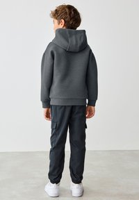 Copil cu păr scurt și castaniu, purtând un hanorac texturat gri închis, pantaloni cargo negri și adidași albi, stând cu fața spre departe pe un podea deschisă la culoare.