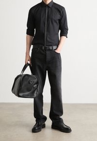 Camisa negra de botones con cremallera vertical, jeans oscuros, cinturón de cuero negro, bolsa de deporte negra con asa y zapatos negros con suela plana.