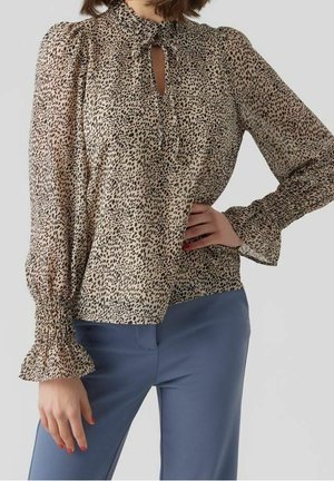 Femme portant un chemisier beige à manches longues avec imprimé animalier noir et poignets volantés, associé à un pantalon bleu taille haute.