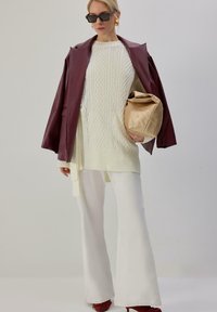 Pull en laine crème à torsades, manteau en cuir bordeaux, pantalon évasé blanc, sac oversize doré et chaussures à talons rouges. Lunettes de soleil noires.