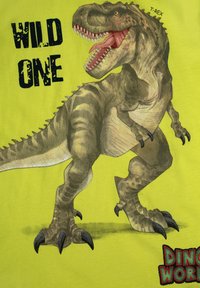 T-shirt verde con una grafica dettagliata di T-Rex, strisce scure, artigli affilati e il testo "WILD ONE" in bold nero, insieme al logo "DINOSAUR WORLD".