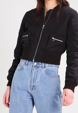 Kvinde iført en cropped sort bomberjakke med lynlåslommer og højtaljede lyseblå jeans, foran en lys baggrund.