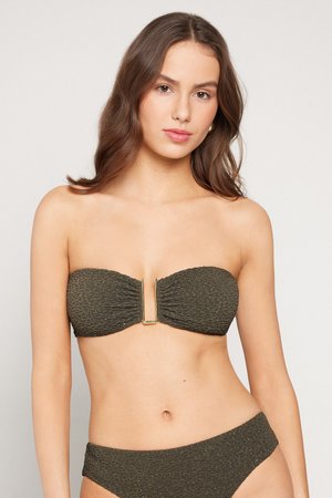 Joven mujer con largo cabello castaño lleva un bikini tipo bandeau texturizado en tono oliva oscuro con acento dorado en la parte superior, posando frente a un fondo claro y sencillo.
