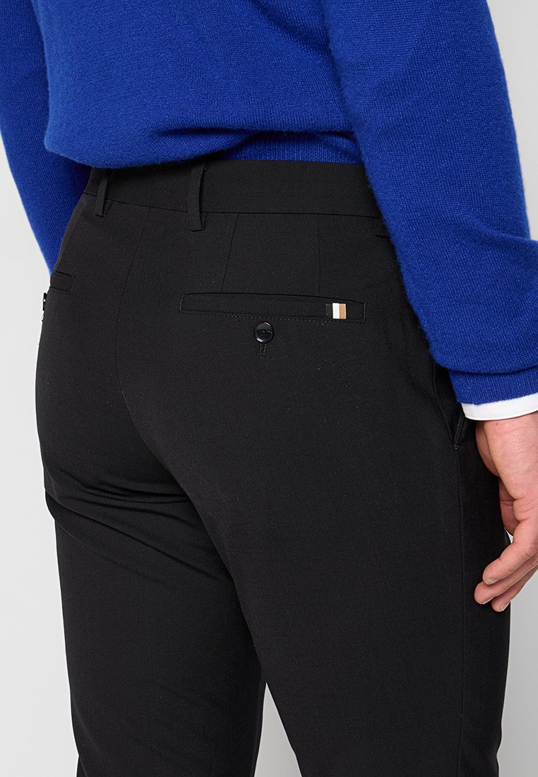 Person mit enganliegenden schwarzen Hosen mit aufgesetzter Gesäßtasche und blauem Pullover, gesehen von der Taille abwärts, rechter Hand sichtbar an der Seite.