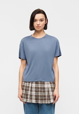 Kvinde med kort mørkt hår iført en blå kortærmet top over et brunt ternet skørt og blå jeans, står foran en ensfarvet lys baggrund.