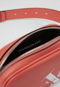 Borsa a tracolla in finta pelle corallo con cerniera nera. L'interno presenta il logo "CALVIN KLEIN JEANS". Design piatto con tracolla regolabile.