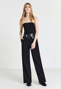 Jumpsuit nero senza spalline con design a gamba larga, dotato di una cintura nera con borchie e texture liscia. Il modello posa con le mani nelle tasche.