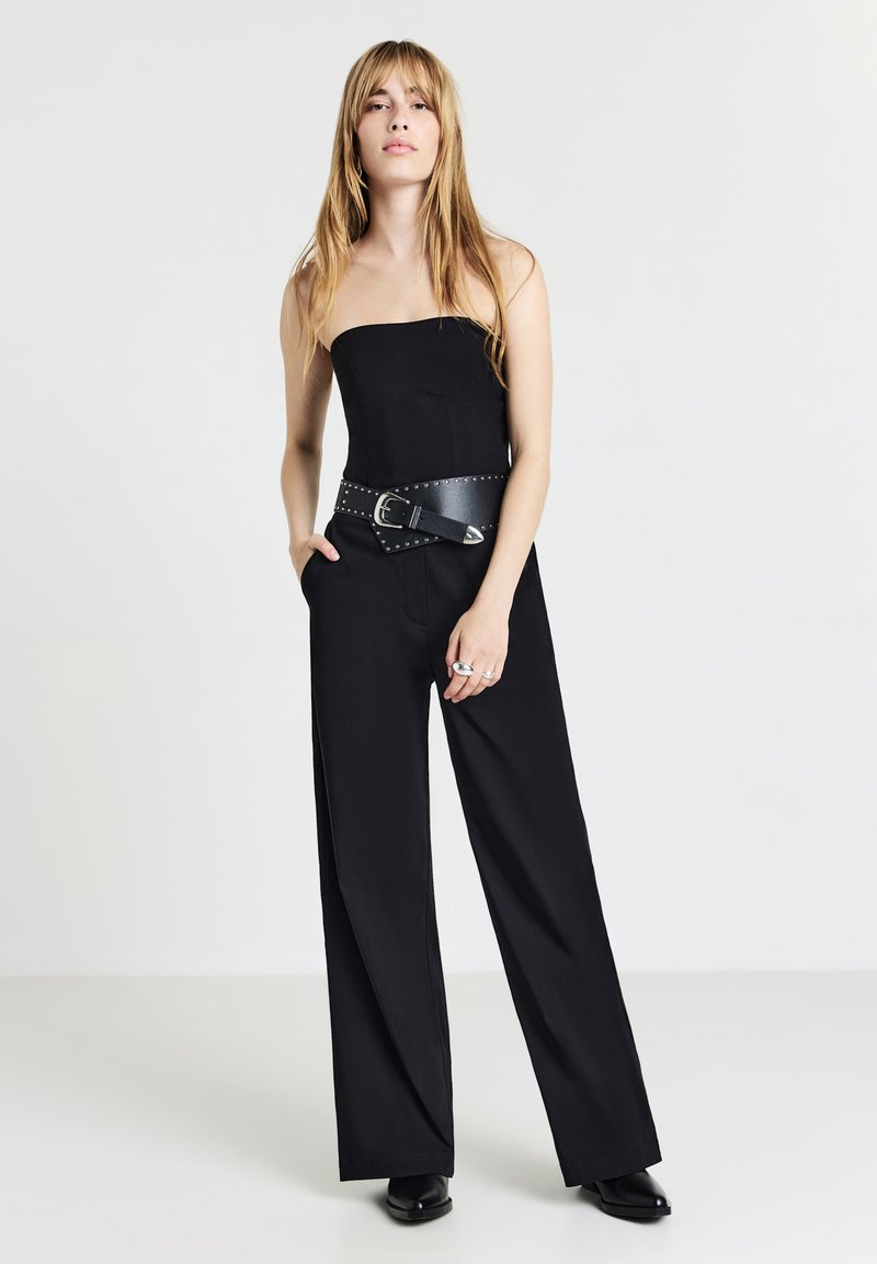 Jumpsuit nero senza spalline con design a gamba larga, dotato di una cintura nera con borchie e texture liscia. Il modello posa con le mani nelle tasche.