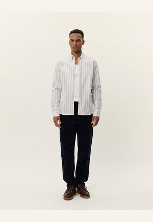 Button-up shirt with vertical light and dark stripes, white background. Paired with dark navy pants. Model wears a white undershirt. Brown shoes.