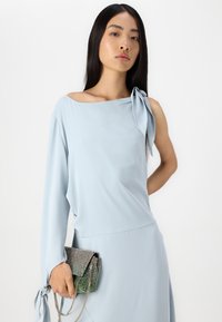 Ανοικτό μπλε ασύμμετρο τοπ με άνοιγμα ώμου και φιόγκο λεπτομέρεια; λείο ύφασμα. Το άτομο κρατά μια λαμπερή τσάντα φάκελο με διαβαθμίσεις χρωμάτων και αλυσίδα.