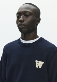 Pull en maille marine à col rond, avec un col côtelé texturé et un logo blanc en forme de lettre "W" sur le côté gauche de la poitrine. Fond uni.