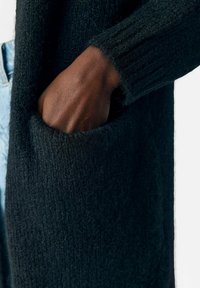Svart stickad cardigan med ribbad mudd och sidoficka, med en mjuk, texturerad finish. Tyget verkar varmt och tjockt.