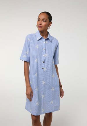 Mujer con vestido camisero de manga corta de color azul claro con patrones florales blancos, de pie con las manos relajadas, mirando ligeramente hacia su izquierda.