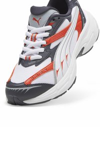 Kicks Puma v bílé síťovině s šedými a oranžovými syntetickými akcenty. Mezi funkce patří bílé tkaničky, polstrovaná podrážka a výrazné branding.