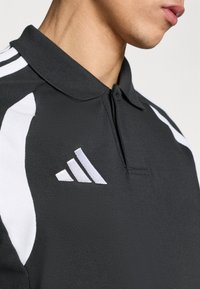 Μαύρο πόλο πουκάμισο με γιακά, λευκές πλευρικές λεπτομέρειες και κεντημένο λευκό λογότυπο adidas στο στήθος. Υφασμάτινη υφή.