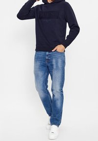 Sudadera azul marino con el logotipo "GUESS" texturizado, combinada con jeans de color azul claro y zapatillas blancas, con un ajuste relajado y diseño casual.