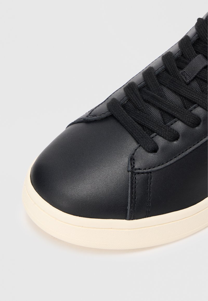 Black leather sneaker with a rounded toe, lace-up design, and a cream rubber sole. Smooth texture with contrasting stitching detail.
