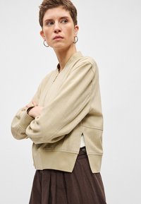 Μπεζ μπουφάν τύπου bomber με λεία υφή, κλείσιμο με φερμουάρ και ελαστικές μανσέτες. Διαθέτει πλαϊνές σχισμές για άνεση και χαλαρή εφαρμογή.