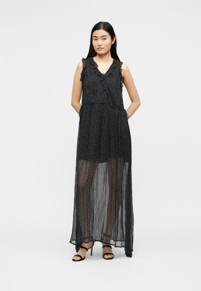 Γυναίκα με μαύρο αμάνικο μάξι φόρεμα με λευκές πουά και διάφανο ύφασμα από το μέσο του μηρού και κάτω, φοράει μαύρα πέδιλα με λουριά, στέκεται με τα χέρια πίσω.