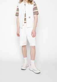 Weißer Denim-Westen mit Reißverschluss, gestreiftes Kurzarmhemd in Beige und Rost, weiße Shorts und weiße High-Top-Sneaker.