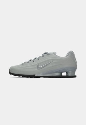 Sānu skats uz gaiši pelēku Nike sporta apavu ar šņorēm, swoosh logo un caurspīdīgu amortizējošu zoli uz vienkārša fona.