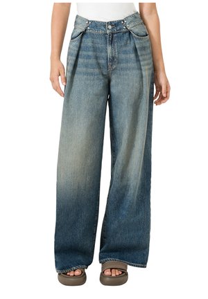 Spodnie dżinsowe z szerokimi nogawkami w wyblakłym niebieskim kolorze, z plisami i metalowymi guzikami, noszone z beżowymi sandałami na otwartym palcu i białym topem.