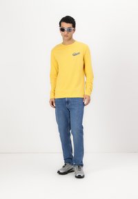 Gelb langärmliges Hemd mit Logo, blaue Jeans und graue Sneaker. Accessoires umfassen übergroße Sonnenbrillen. Einfaches Design, ohne Muster.