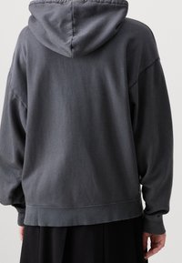 Gray hoodie with a loose fit, kangaroo pocket, and ribbed cuffs. Features a large hood and soft cotton texture, worn over a black skirt.