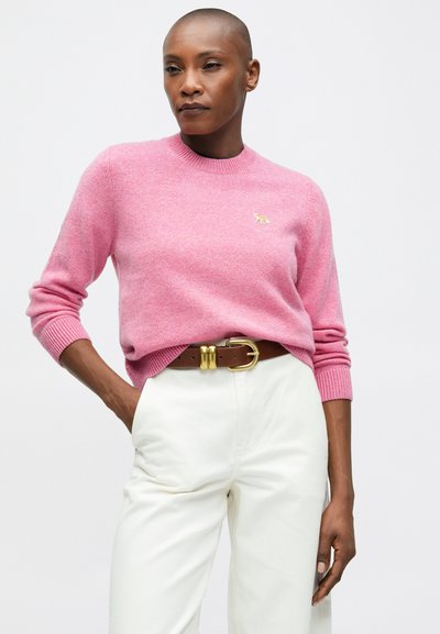 Kobieta w różowym swetrze z małym haftowanym logo, w białych, wysokich spodniach i brązowym pasku ze złotą klamrą, pozująca z ręką w kieszeni.