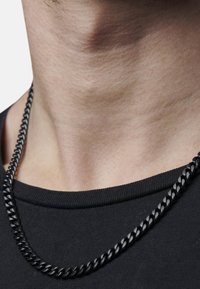 Sort metal kædedesign med en snoet struktur, der hviler på en bar hals, parret med en sort t-shirt med rund hals. Glat tekstur synlig.