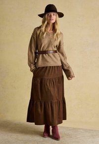 Brązowy sweter dzianinowy z dekoltem w serek, brązowa, warstwowa maxi spódnica, bordowe botki sięgające kostki oraz szerokiego ronda brązowy kapelusz.