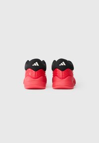 Des chaussures de sport rouges avec des cols rembourrés noirs, un design de talon ondulé et un logo à trois rayures blanches, vues de l'arrière sur un fond uni.