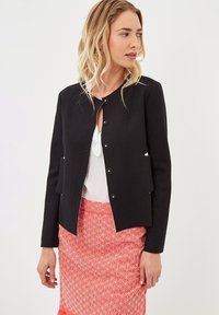 Chaqueta negra entallada con escote redondo y cierre frontal de botones, combinada con una falda coral con un patrón geométrico blanco.