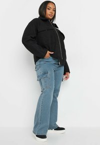 Chaqueta negra con cremallera y diseño estructurado, equipada con dos bolsillos en el pecho, combinada con pantalones cargo de color azul claro que cuentan con bolsillos laterales y piernas acampanadas.
