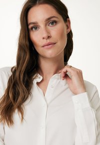 Camicia bianca con colletto classico, bottoni in perla e maniche lunghe. Tessuto liscio con una leggera lucentezza, progettata per una vestibilità su misura.