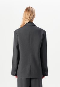 Person mit langen blonden Haaren, die eine übergroße dunkelgraue Anzugjacke und passende Hose trägt, steht mit dem Rücken zu einem schlichten weißen Hintergrund.