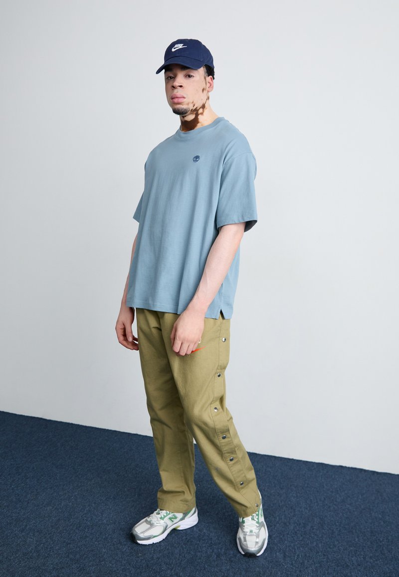 T-shirt en coton bleu clair avec un petit logo, associée à un pantalon cargo vert olive avec des boutons-pression sur les côtés, complétée par des baskets grises.