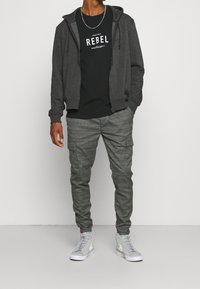 Grå zip-hoodie över en svart T-shirt med texten "REBEL", kombinerad med grå rutiga cargobyxor och ljusgrå sneakers.