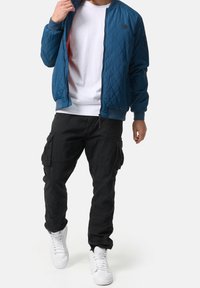 Man die een blauwe gewatteerde jas draagt over een wit T-shirt, zwarte cargobroek en witte sneakers, lopend tegen een effen witte achtergrond.