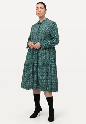 Teal jurk met een knoopsluiting kraag, lange mouwen, gelaagd ontwerp en een gestructureerd patroon. Draagt met zwarte haklaarzen tot de enkels.