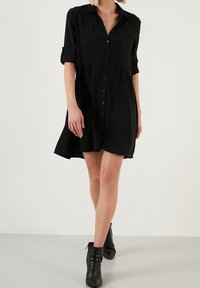 Abito nero a camicia con chiusura a bottoni sul davanti, colletto e maniche arrotolate. Realizzato in tessuto leggero, presenta una vestibilità ampia e un orlo svasato.